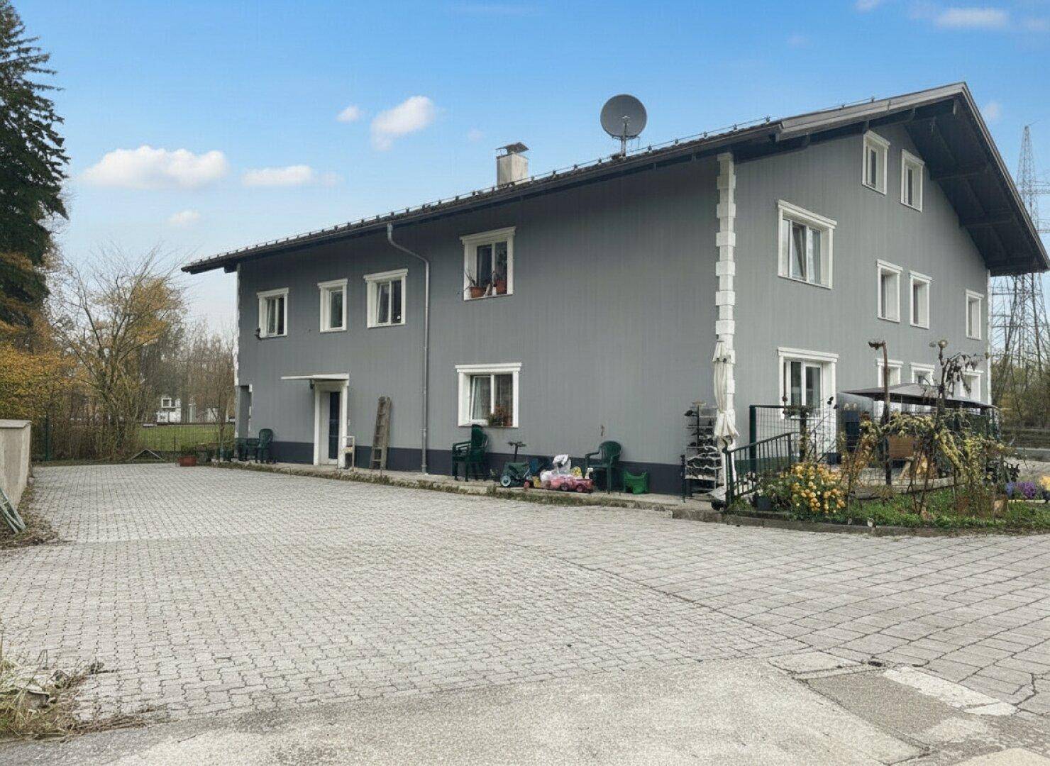 Seitenansicht des grauen Gebäudes mit großem gepflasterten Parkplatz und Gartenbereich.
