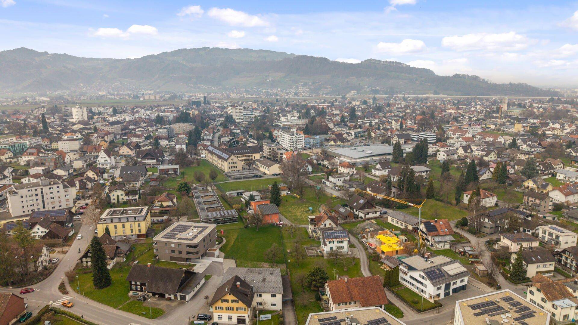 Panoramablick aus der Luft auf eine Stadt mit Wohn- und Gewerbegebieten vor einer bewaldeten Bergkette.