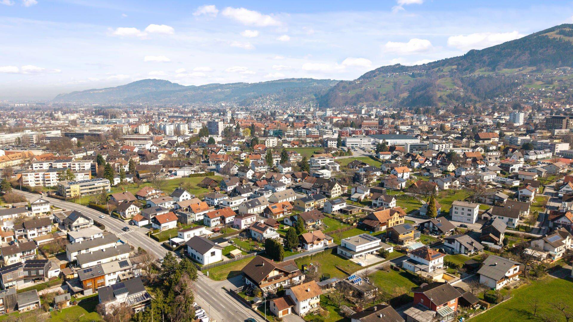 Luftaufnahme einer Stadt mit Wohnsiedlungen, umgeben von grünen Hügeln unter blauem Himmel.