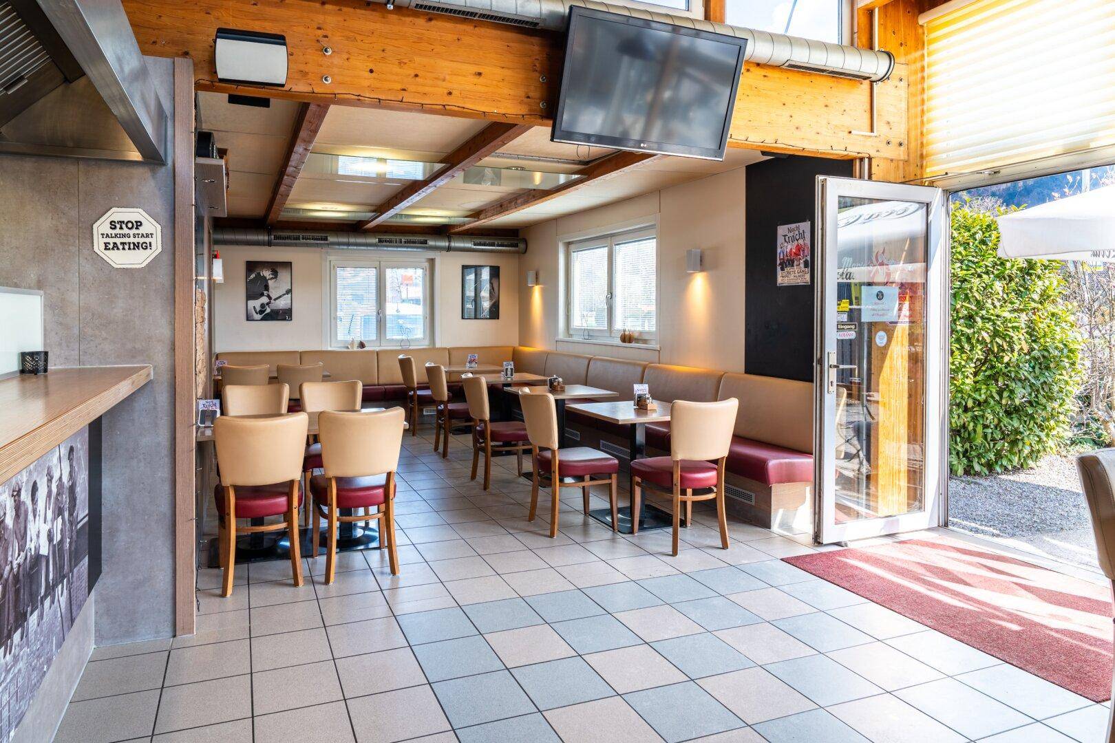 Heller Restaurantbereich mit Fliesenboden, Holzbalken und bequemen Sitzbänken entlang der Fensterfront.