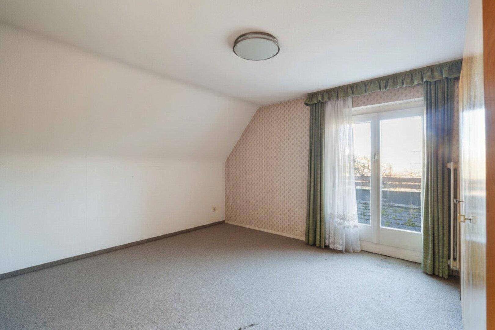 Leeres Zimmer mit Teppichboden, schräger Decke und Zugang zu einem Außenbereich.