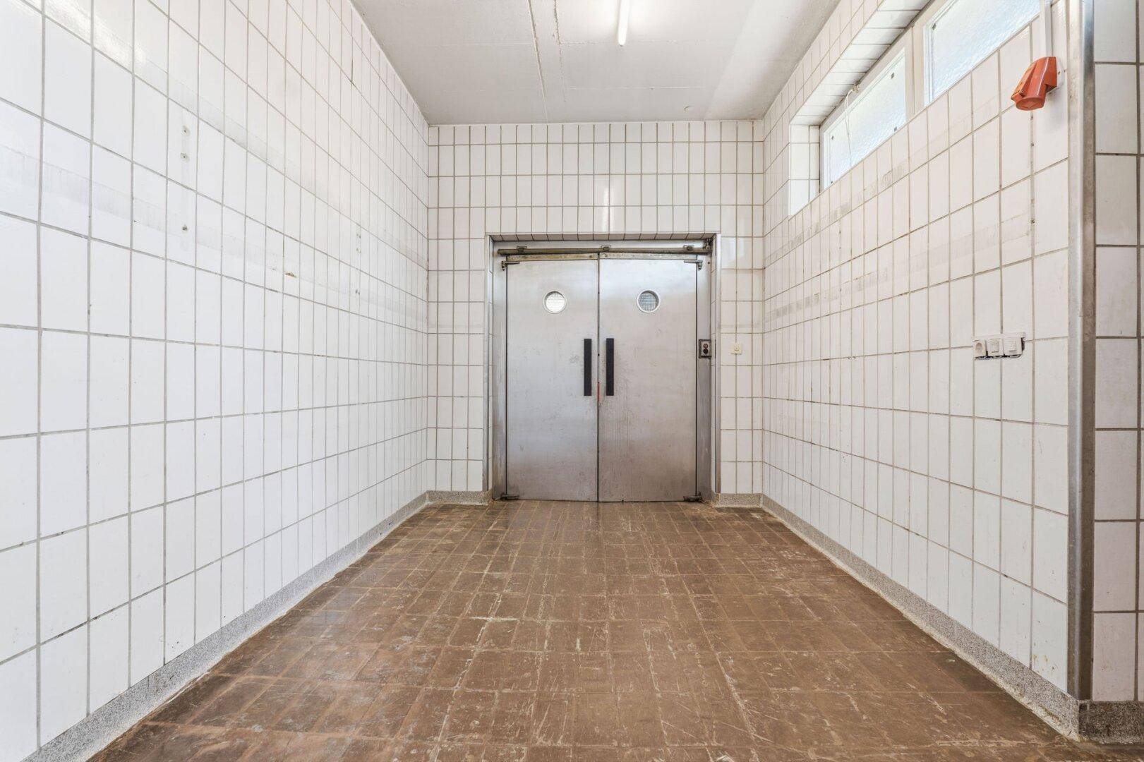Langer Flur mit gefliesten Wänden und großen Metalltüren, die zu einem Aufzug führen.