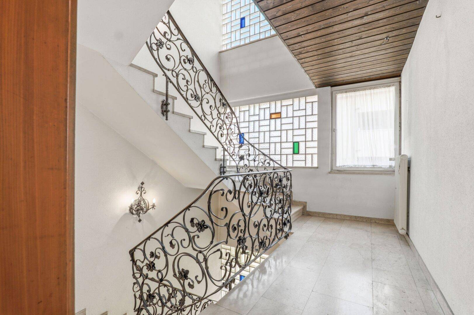 Elegantes Treppenhaus mit schmiedeeisernem Geländer und farbigen Glasbausteinen, das viel Licht hereinlässt.