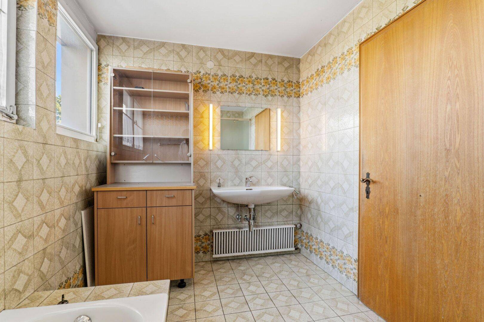 Badezimmer mit gemusterten Fliesen, Waschbecken, Spiegelschrank und Fenster, im Stil der 80er Jahre.