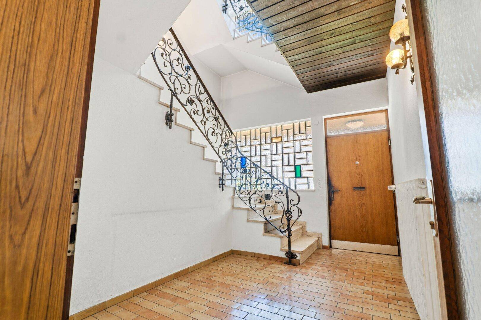 Heller Eingangsbereich mit einer geschwungenen Treppe, schmiedeeisernem Geländer und Fliesenboden.