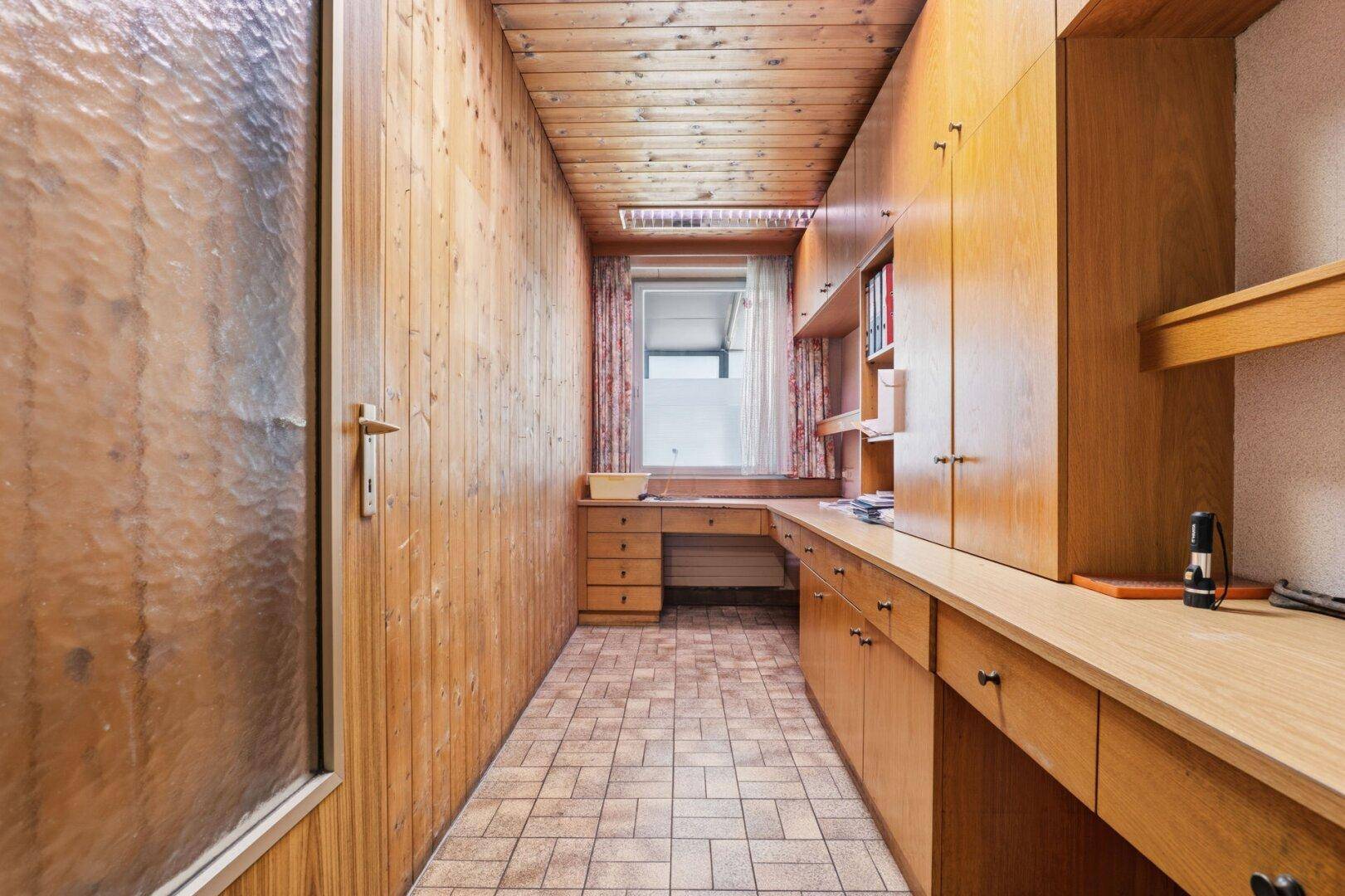 Schmaler Arbeitsraum mit Holzvertäfelung an Wänden und Decke sowie einem integrierten Schreibtisch.