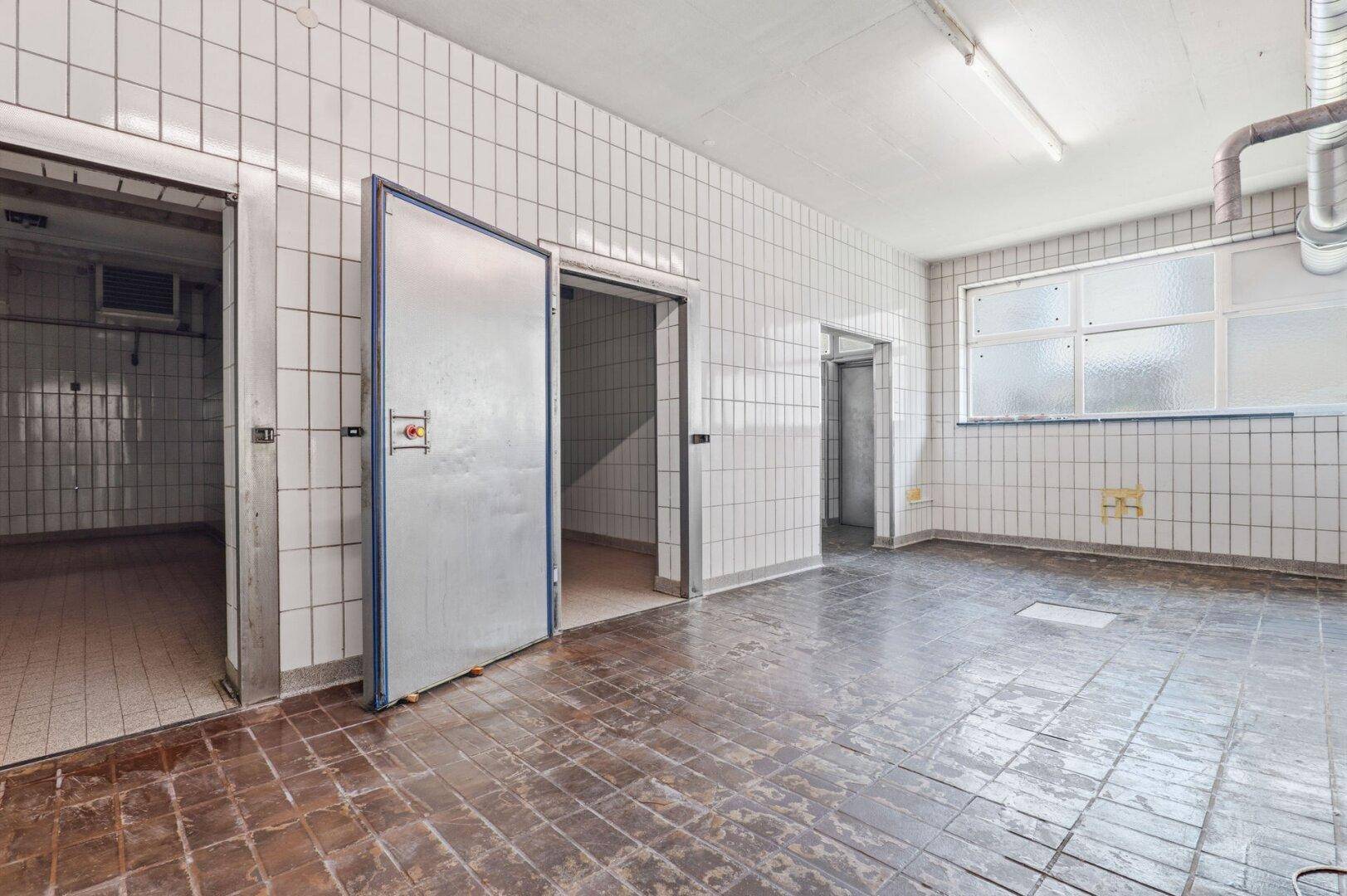 Geräumiger, gefliester Raum mit großer Metalltür und Fenster, geeignet für Lagerung oder gewerbliche Nutzung.