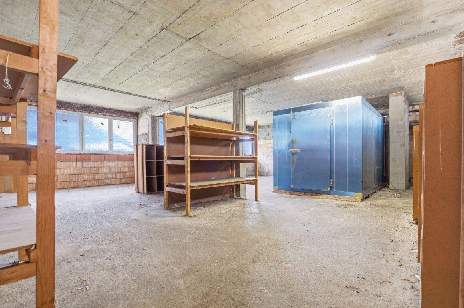 Kellerlagerraum mit Betonboden, Holzregalen und einer großen blauen Kühlzelle.