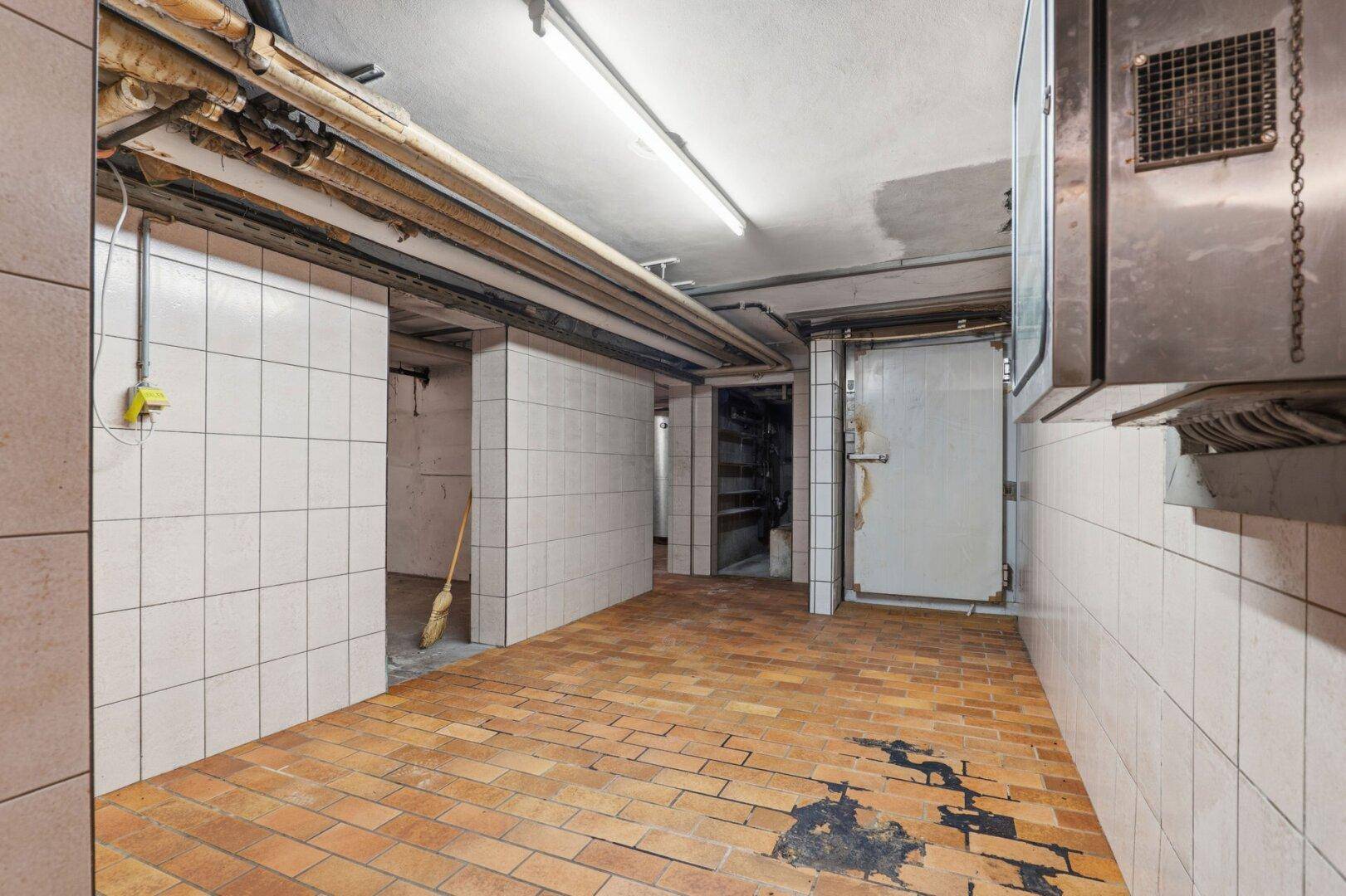 Kellerflur mit weißen Fliesenwänden, Fliesenboden und sichtbaren Rohrleitungen an der Decke.