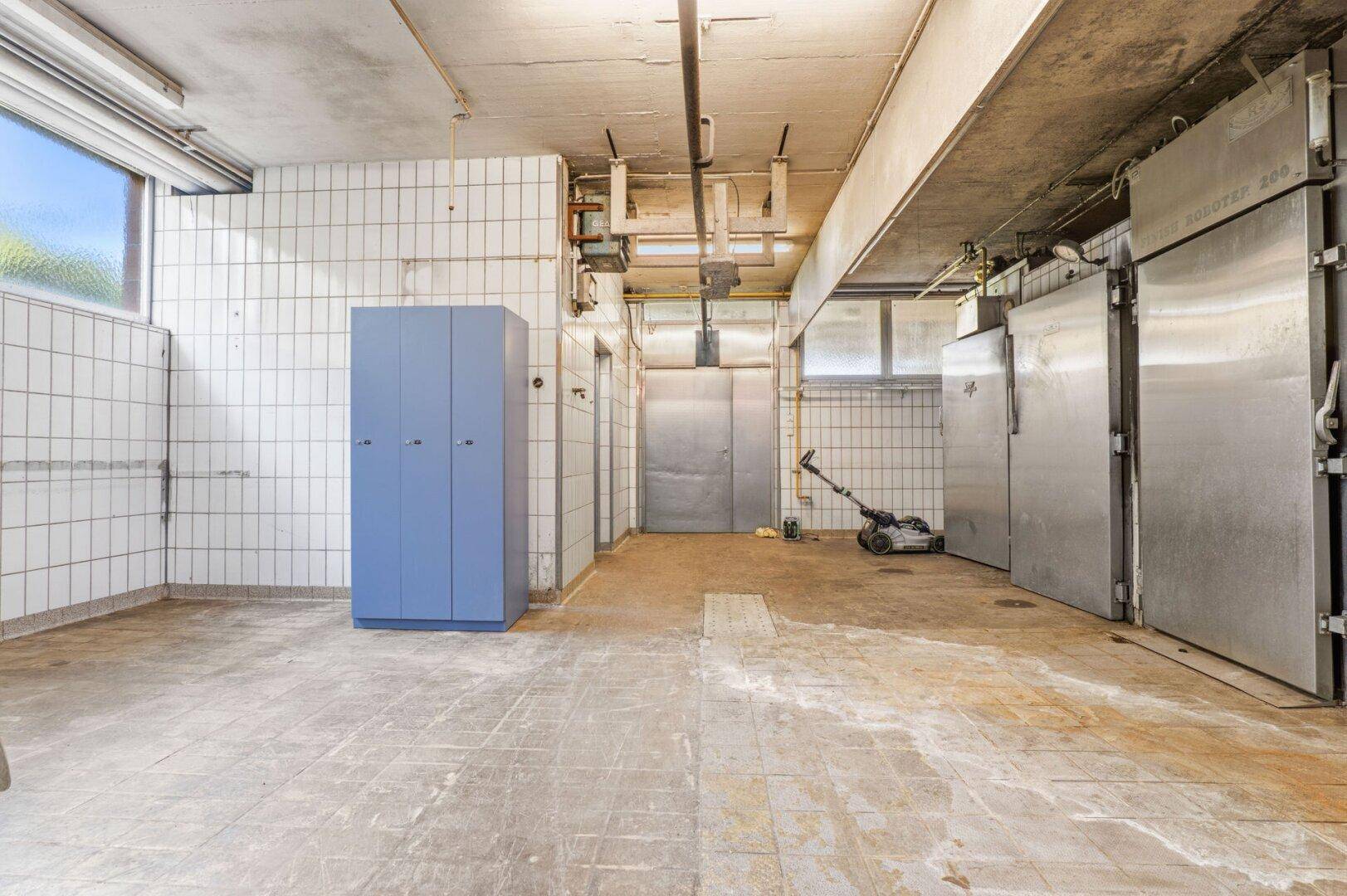 Kellerraum mit weißen Fliesenwänden, Betonboden und mehreren großen Kühlzellen sowie einem Spind.