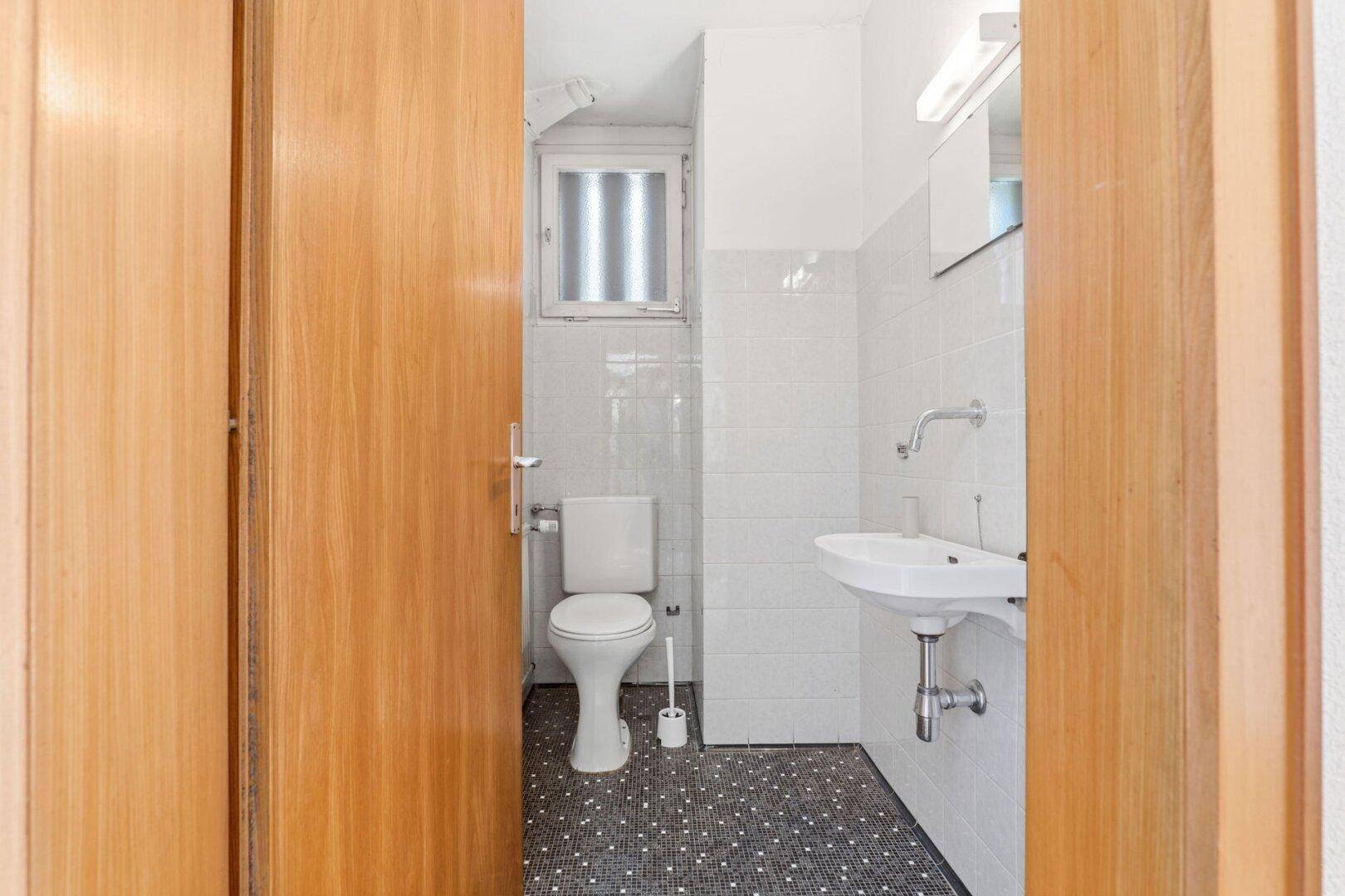 Kleines Gäste-WC mit weißen Fliesenwänden, dunklem Musterboden, Toilette, Waschbecken und Fenster.