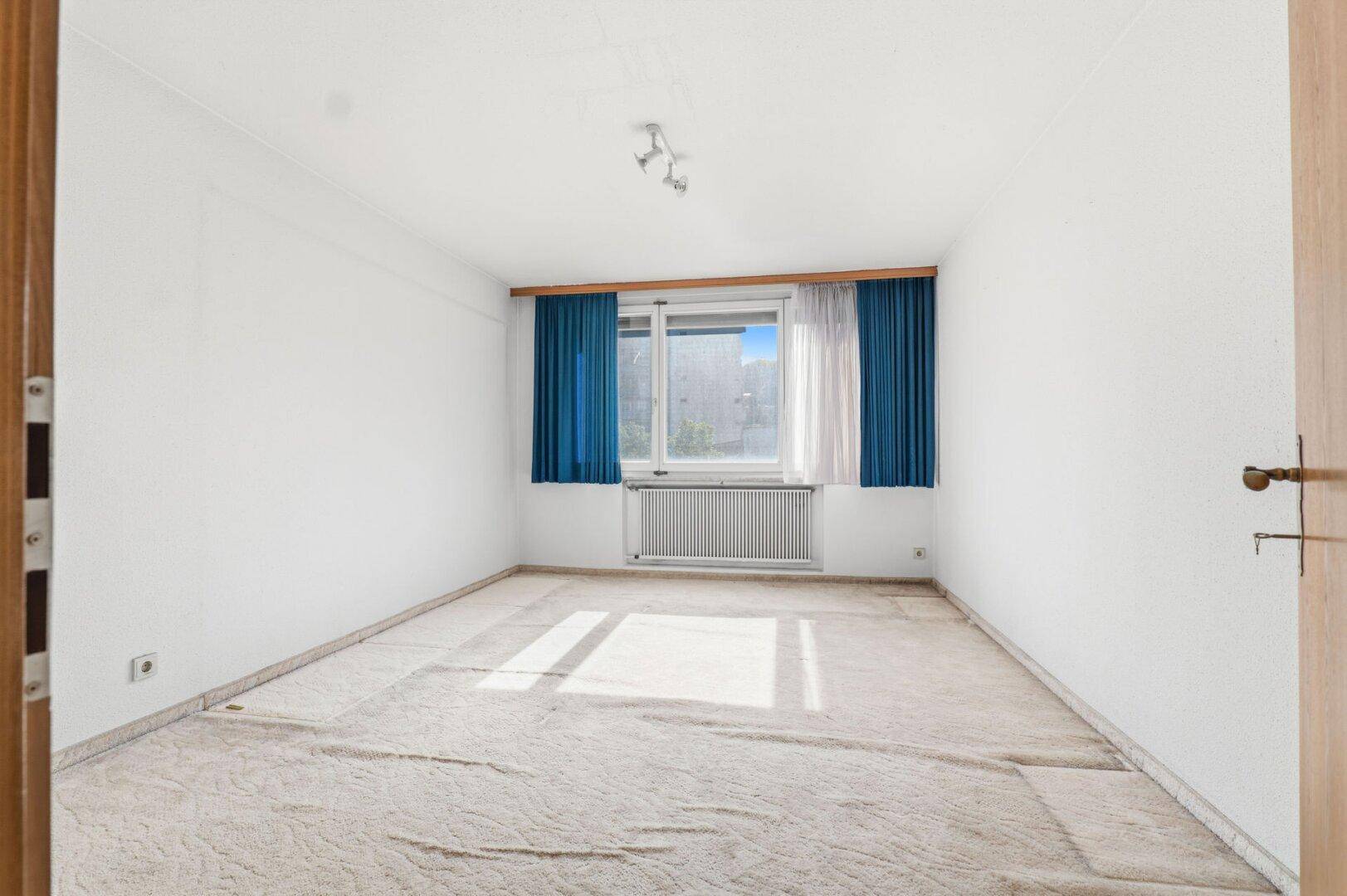 Leeres Schlafzimmer mit Teppichboden, weißen Wänden und einem großen Fenster mit blauen Vorhängen.