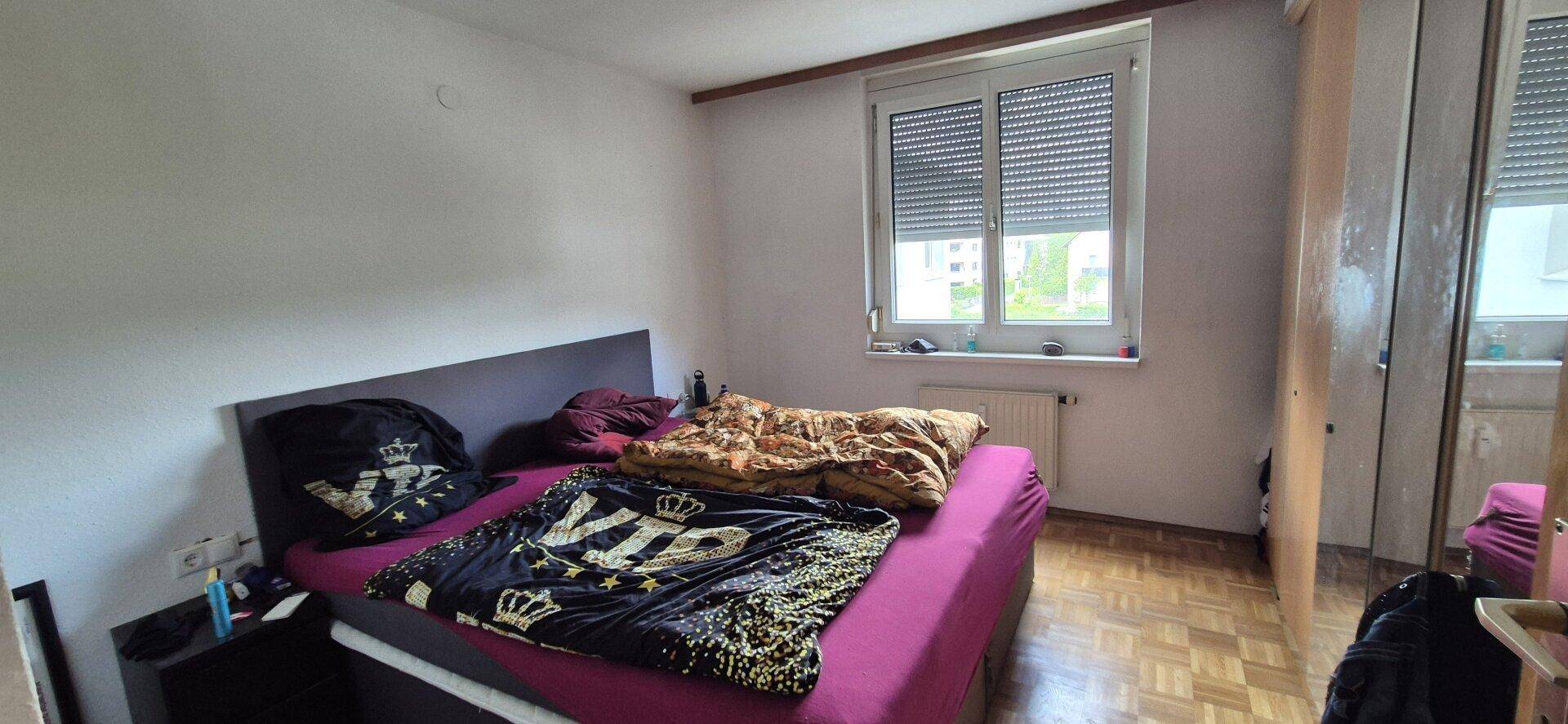 Schlafzimmer mit Bett, Kleiderschrank und Fenster mit Jalousien.