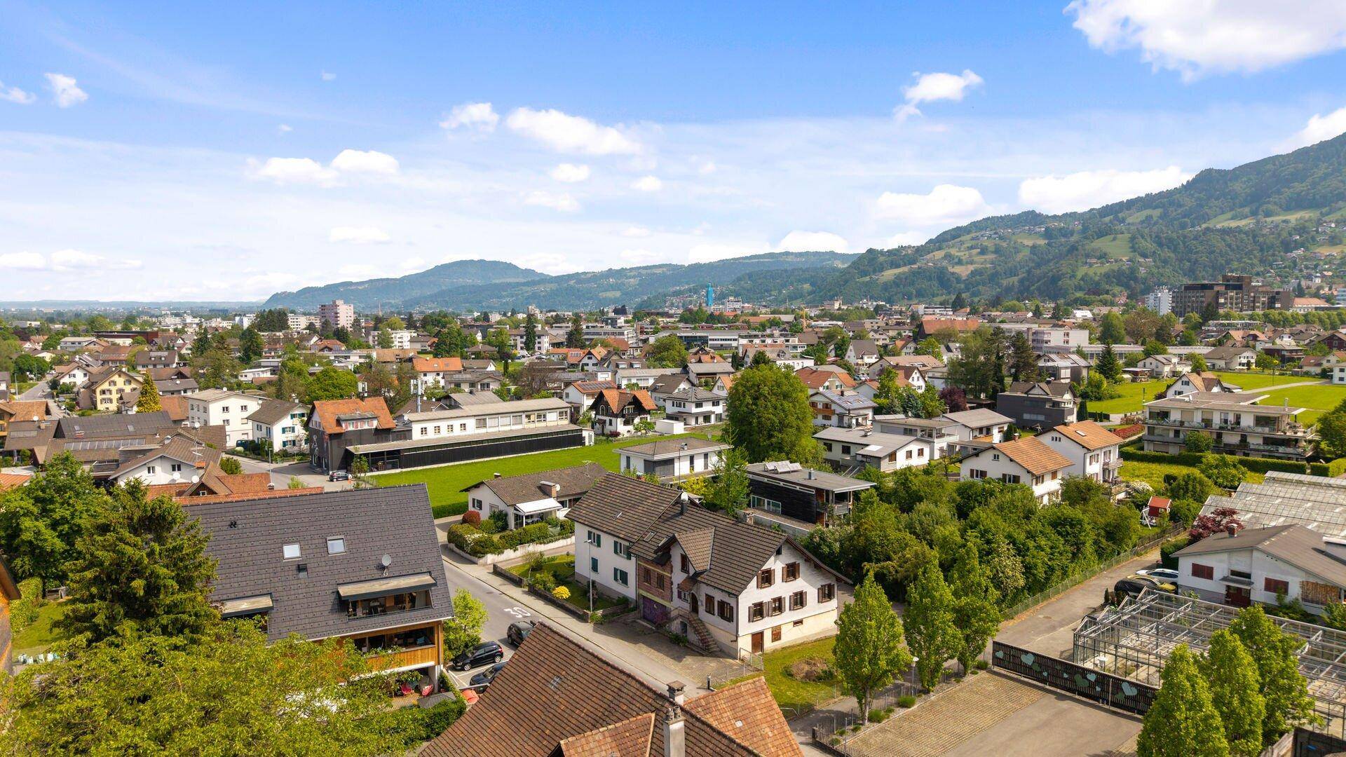 Panoramische Luftaufnahme einer Wohnsiedlung mit grünen Hügeln und Bergen im Hintergrund unter blauem Himmel.