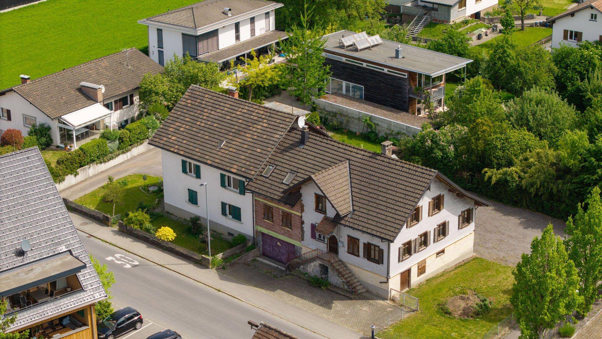 Nahaufnahme eines traditionellen Hauses mit braunem Ziegeldach und grünen Fensterläden, umgeben von Gärten.