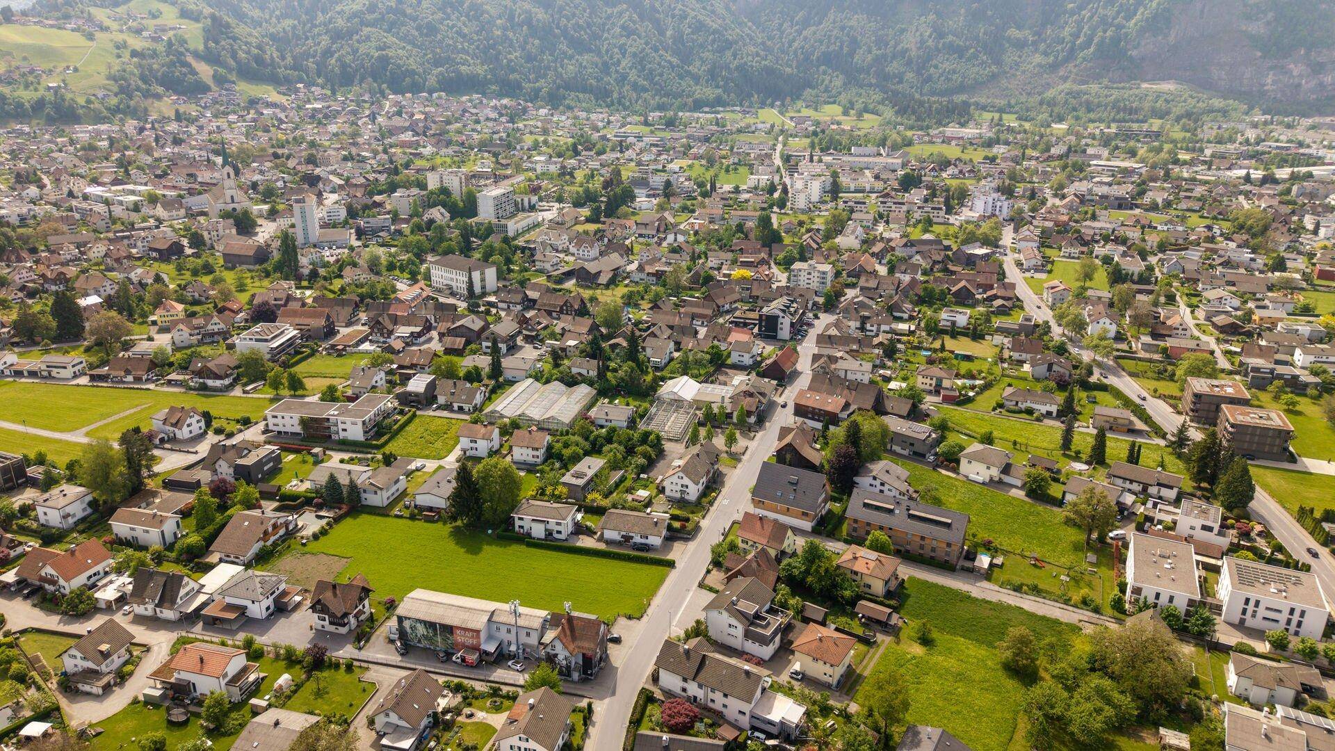 Weitwinkelaufnahme einer städtischen Siedlung mit vielen Häusern und grünen Flächen, umgeben von Bergen.