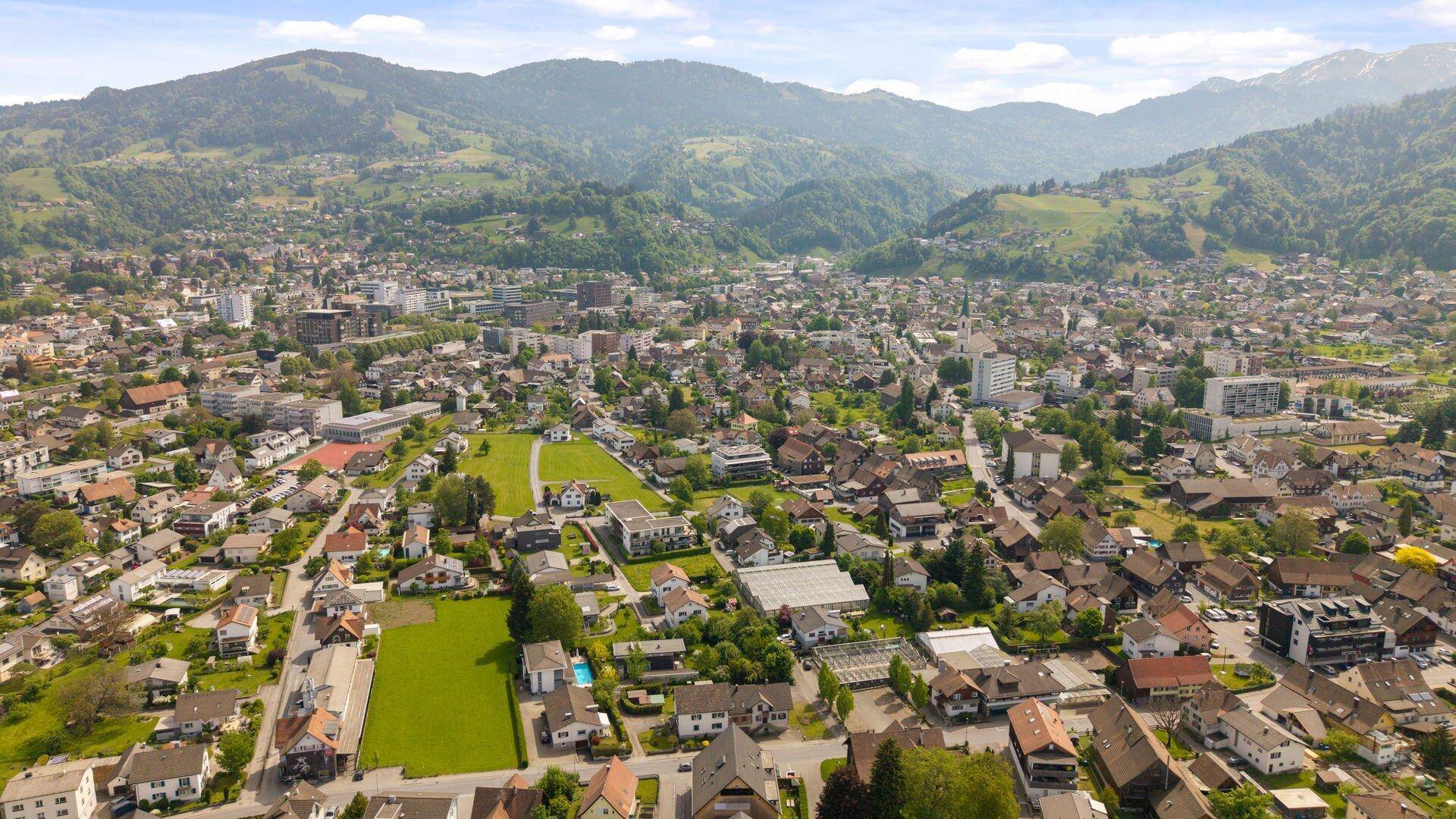 Luftaufnahme einer ausgedehnten Wohngegend mit grünen Wiesen und bewaldeten Bergen im Hintergrund.