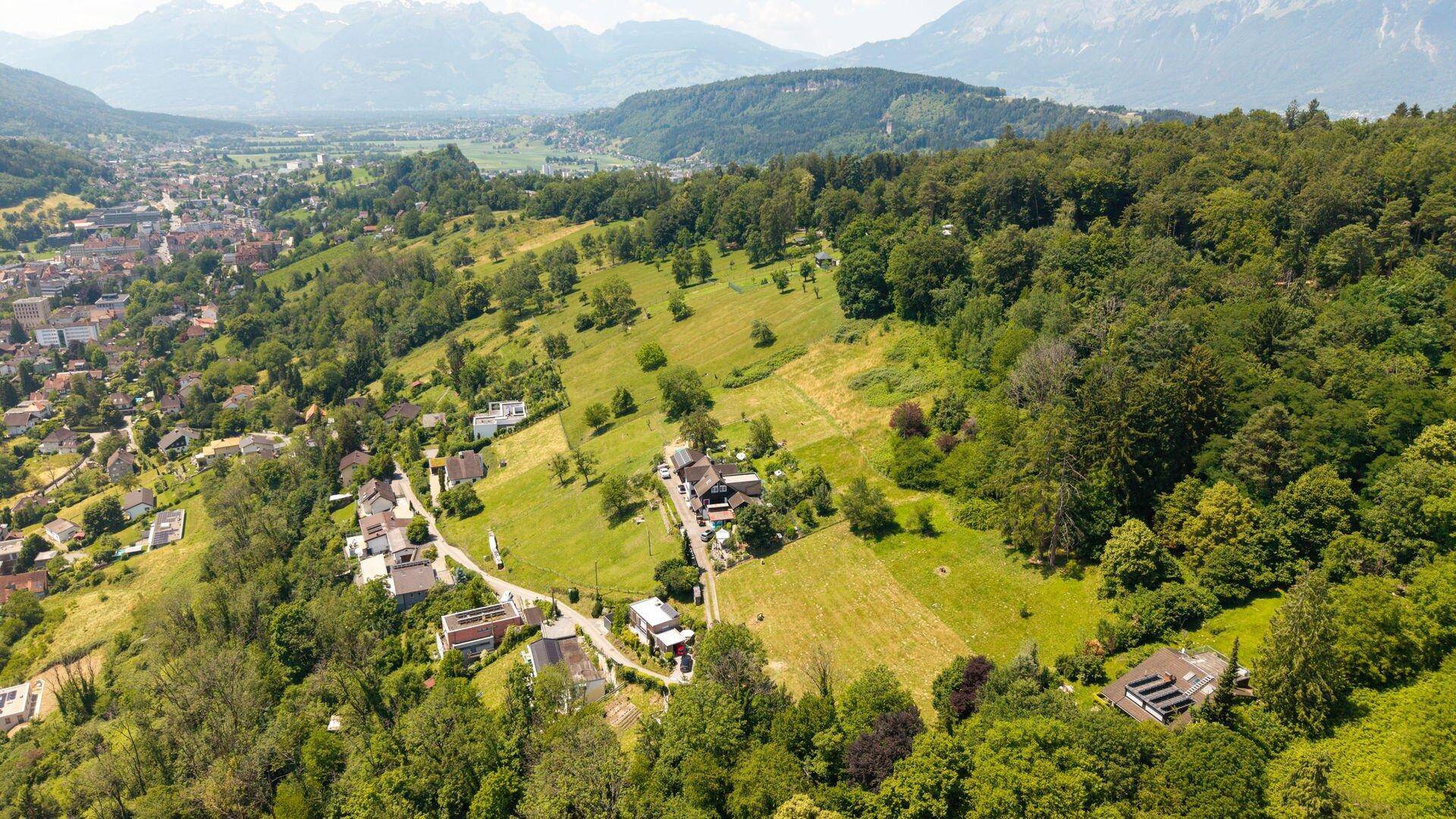 Weitläufige Luftaufnahme einer grünen Hügellandschaft mit verstreuten Häusern und einem Tal mit Stadt im Hintergrund.