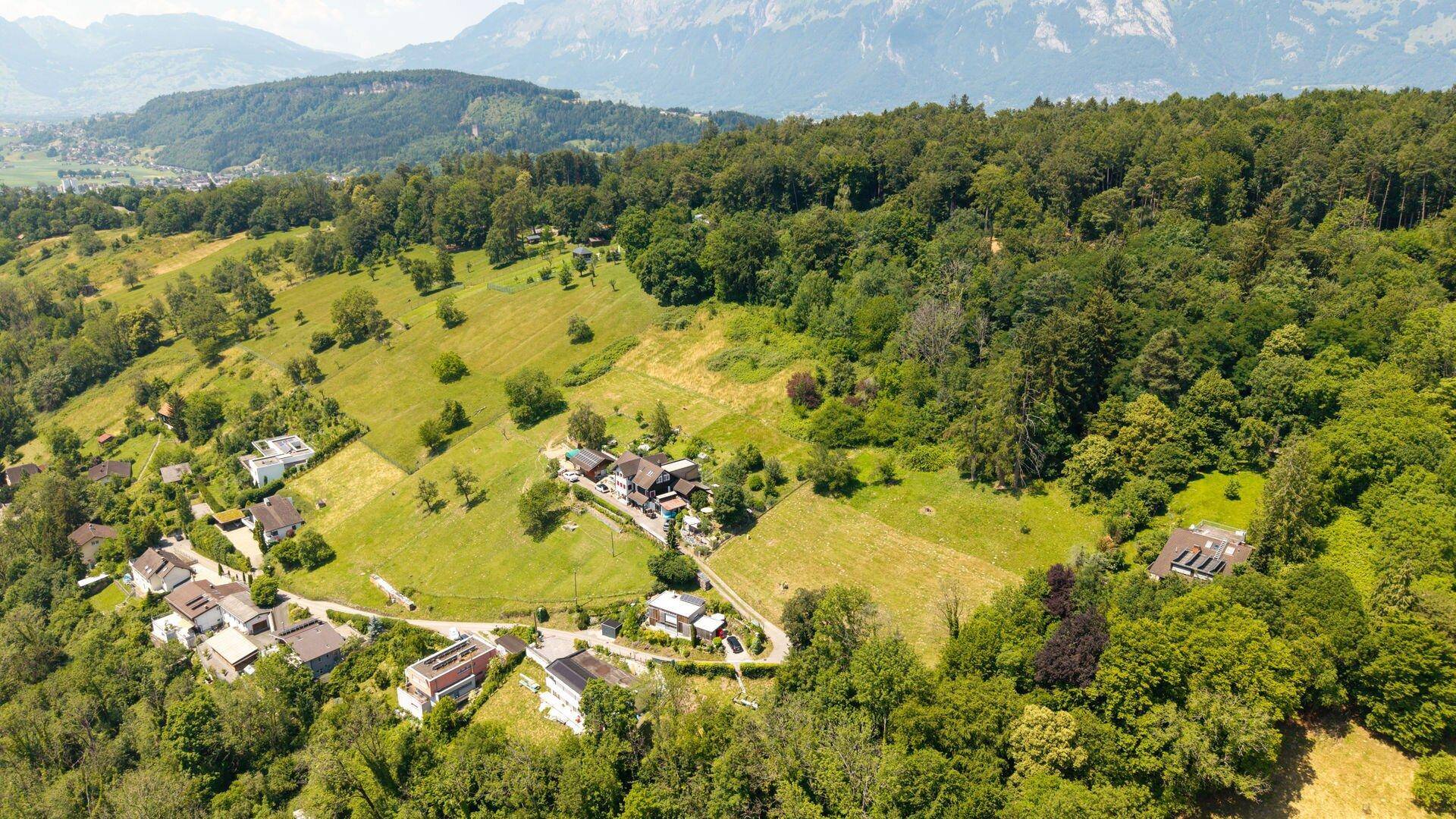 Panoramablick aus der Luft auf eine grüne, hügelige Landschaft mit Häusern, Wäldern und Bergen im Hintergrund.