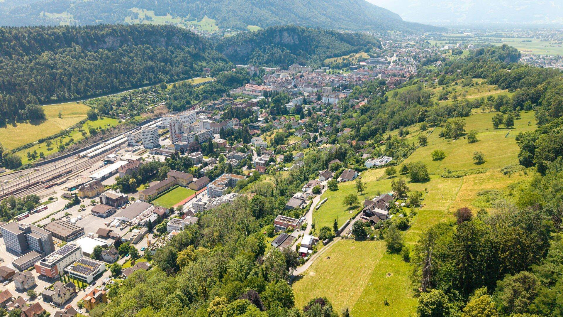Luftaufnahme einer Stadt im Tal, umgeben von grünen Hügeln, Wäldern und einer beeindruckenden Bergkulisse.