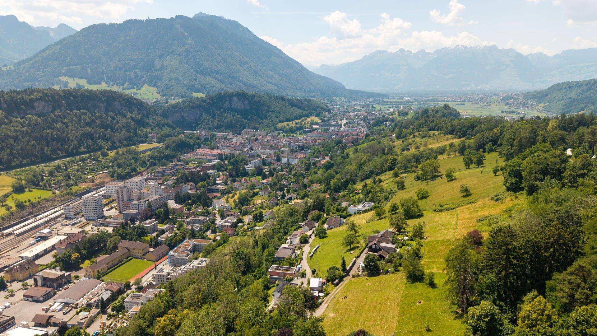 Panoramablick aus der Luft auf eine Stadt im Tal mit Fluss, umgeben von grünen Hügeln und majestätischen Bergen.