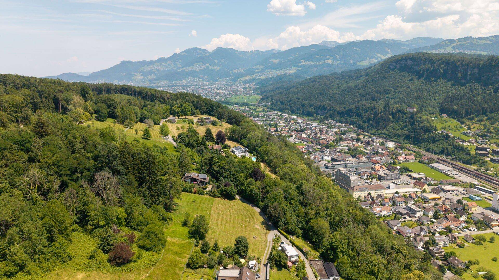 Weitläufige Luftaufnahme einer Stadt im Tal, umgeben von grünen Hügeln, Wäldern und einer weiten Berglandschaft.