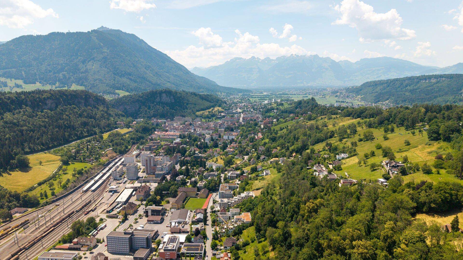 Panoramablick aus der Luft auf eine Stadt im Tal mit Bahngleisen, umgeben von grünen Hügeln und Bergen.