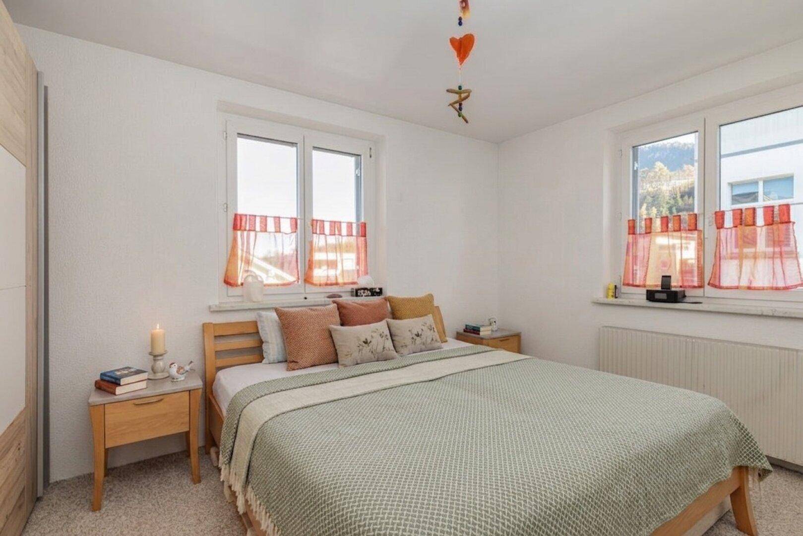 Helles Schlafzimmer mit Doppelbett, Holzmöbeln, Teppichboden und zwei Fenstern, gut gepflegt.