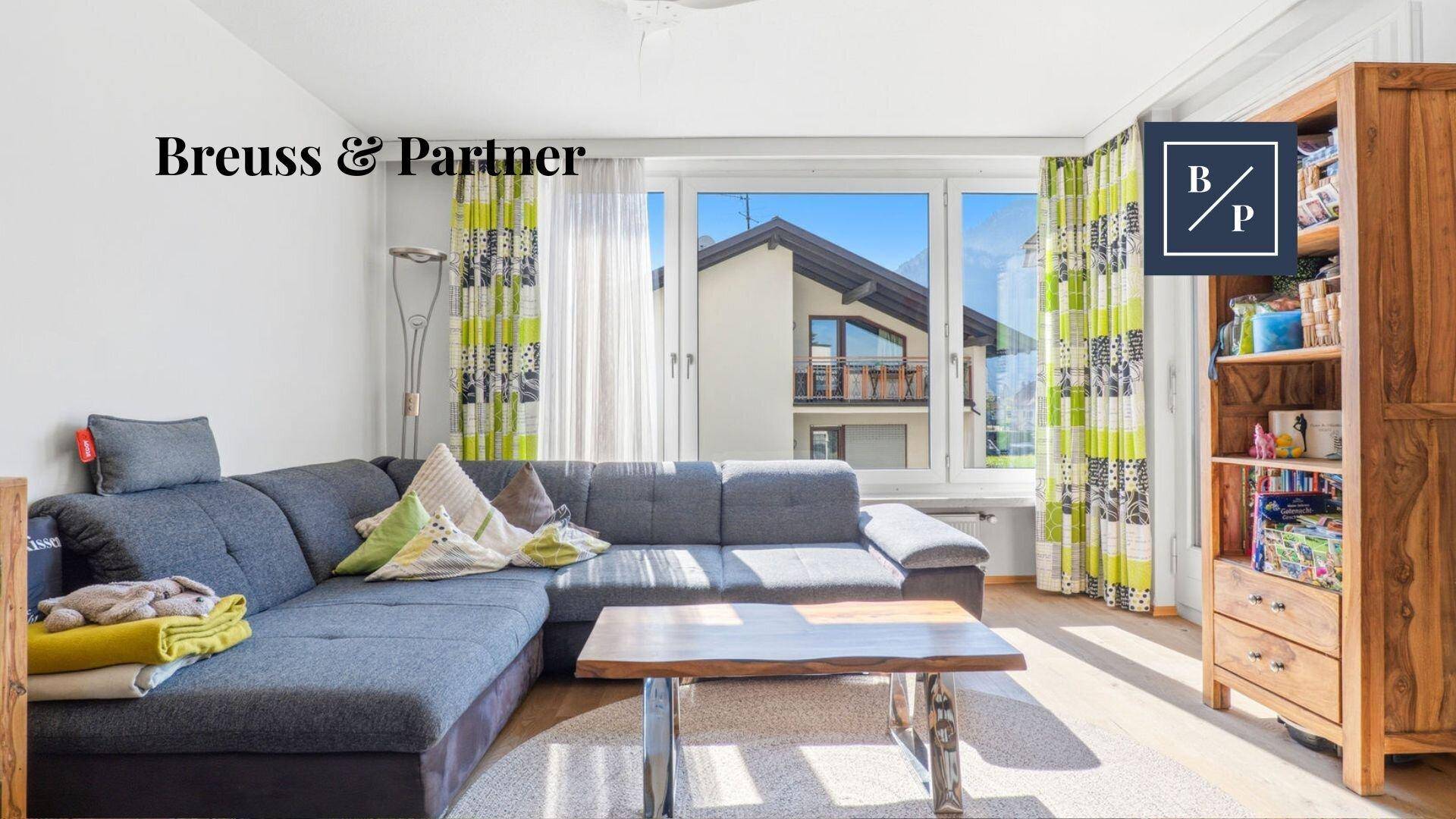 Helles Wohnzimmer mit Ecksofa, Holztisch und Blick auf die umliegenden Häuser.