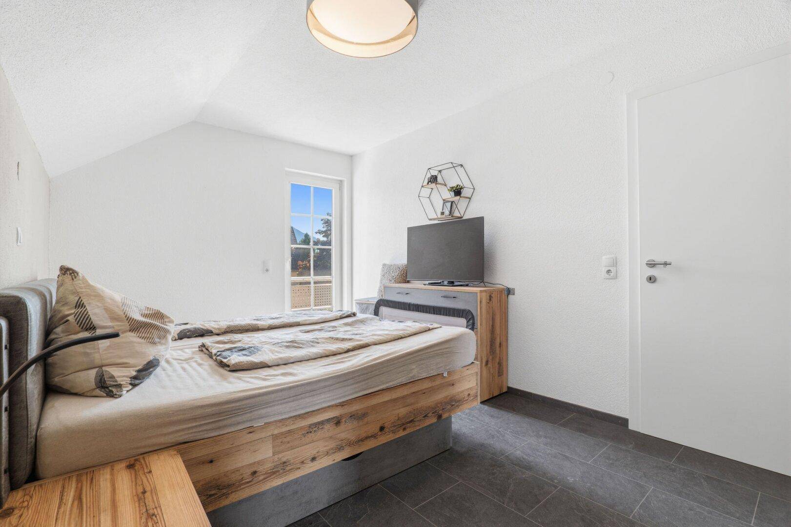 Schlafzimmer mit Doppelbett, dunklen Fliesen und Fenster mit Ausblick.