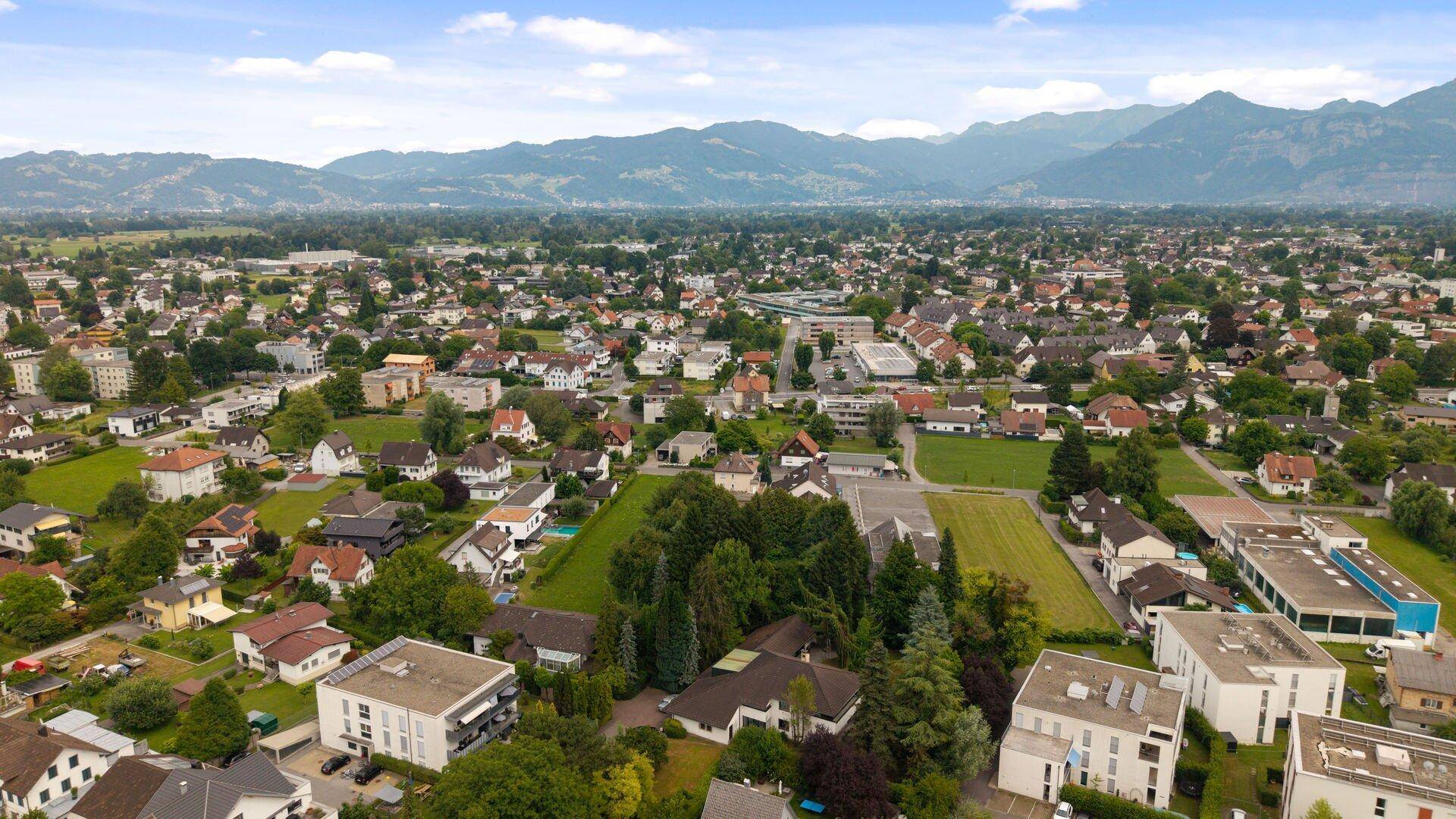 Panoramablick aus der Vogelperspektive auf die Stadt mit grünen Flächen und Bergen im Hintergrund.