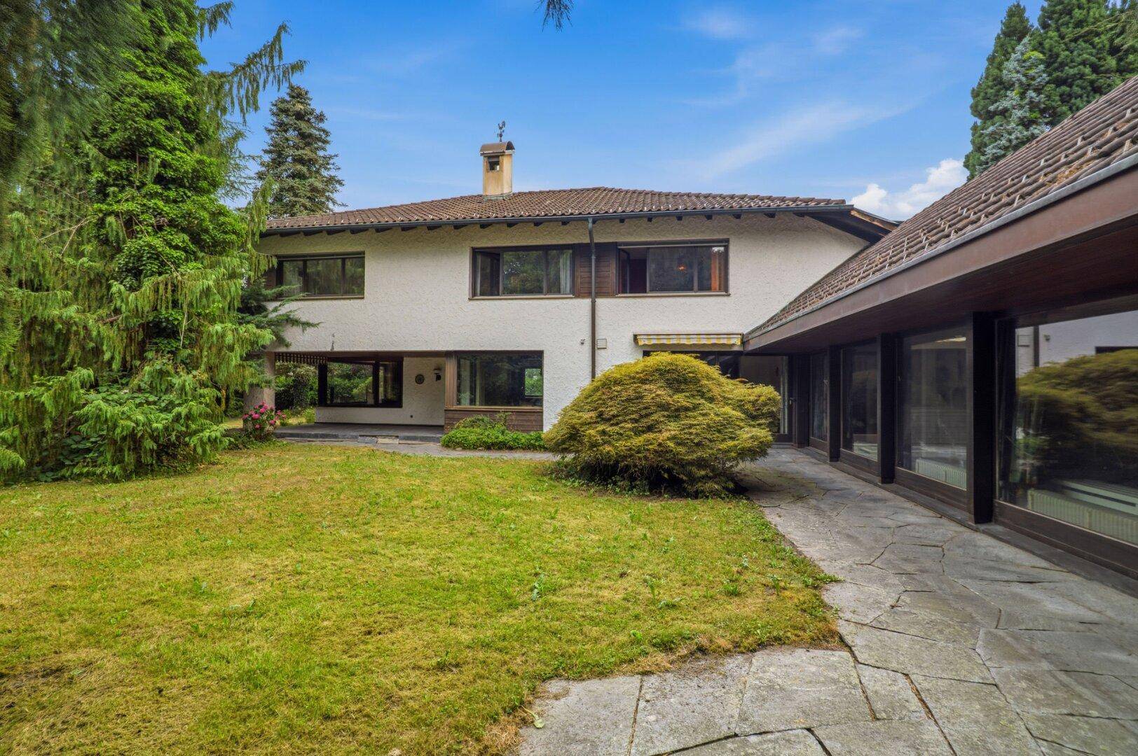 Rückseite des Hauses mit weitläufigem Rasen, Steinterrasse und üppiger Bepflanzung.
