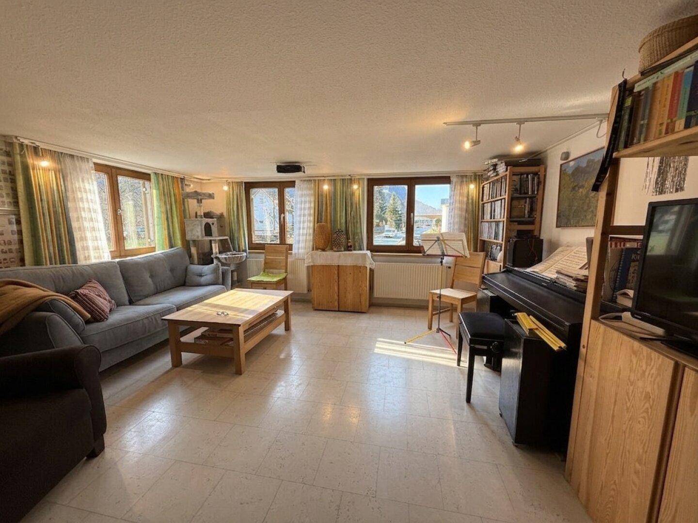 Geräumiges Wohnzimmer mit Sofa, Klavier und Bücherregalen, hell durch mehrere Fenster.