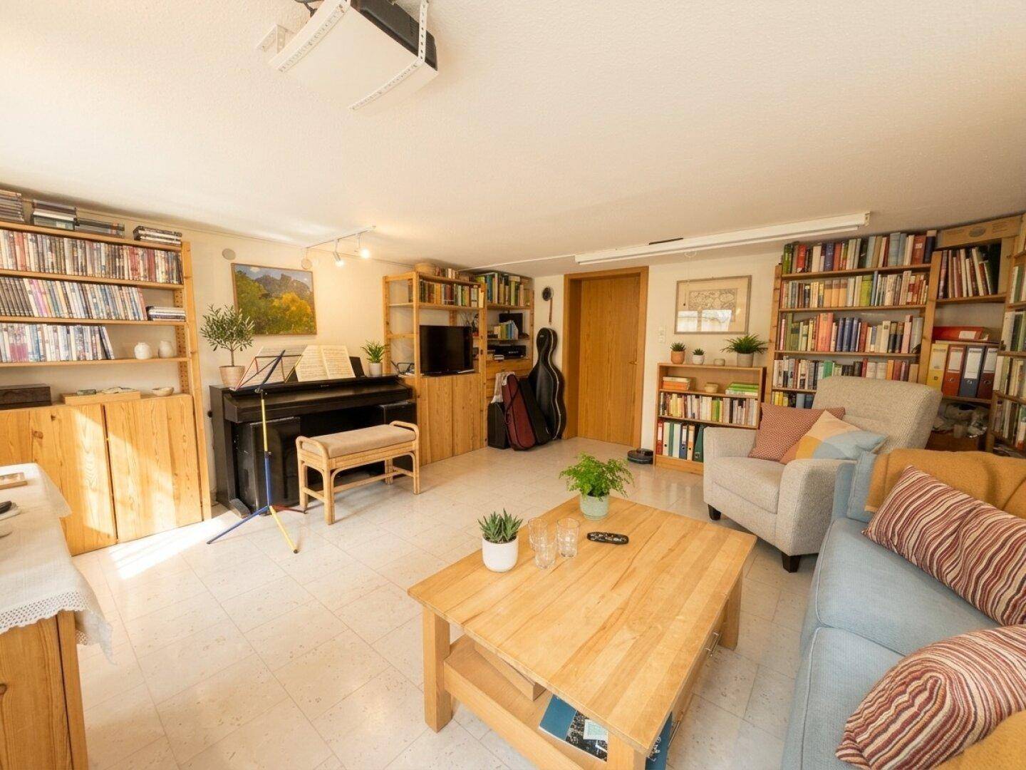 Gemütliches Wohnzimmer mit Sofa, Klavier und umfangreichen Bücherregalen, gut beleuchtet.