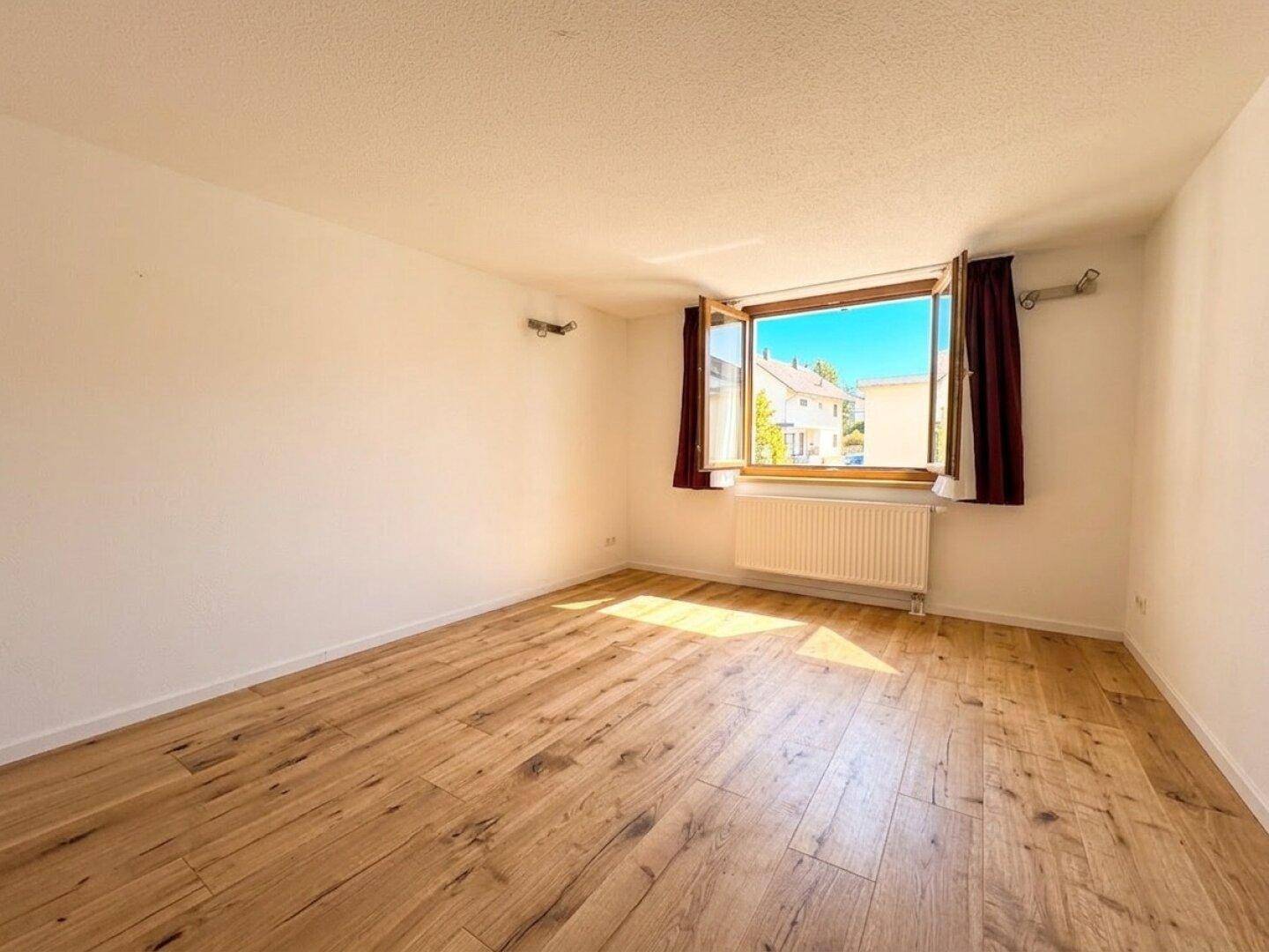 Helles, leeres Zimmer mit Holzboden und großem Fenster, vielseitig nutzbar.