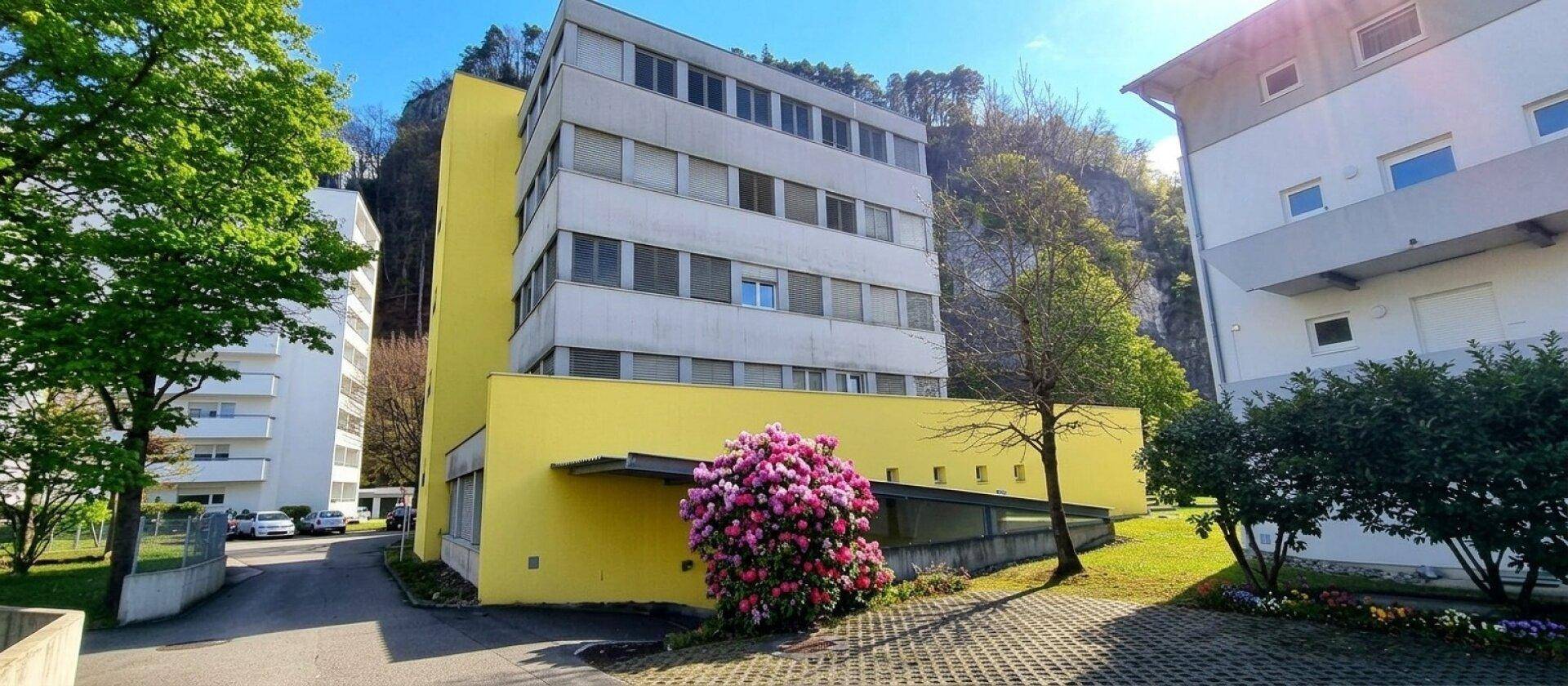 Mehrstöckiges Gebäude mit gelber Fassade und blühenden Sträuchern vor einer felsigen Bergkulisse.