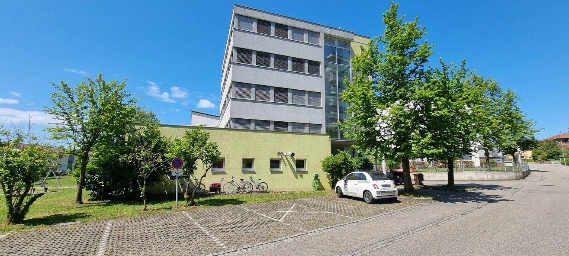 Mehrstöckiges Gebäude mit moderner Architektur, umgeben von Bäumen und einem Parkplatz unter blauem Himmel.