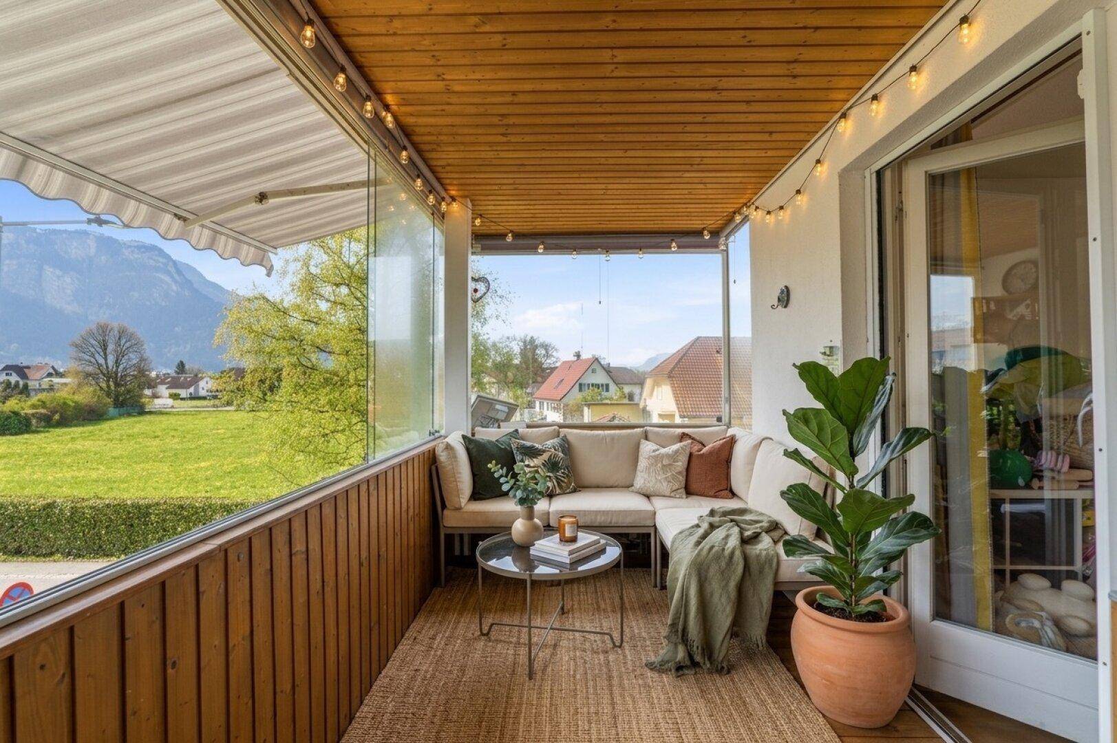 Gemütlicher, verglaster Balkon mit Holzelementen, bequemer Sitzecke und weitem Blick auf die Berge und grüne Landschaft.
