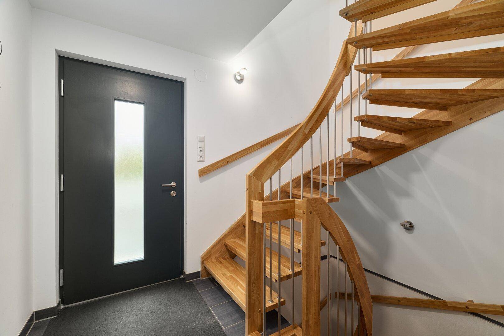 Heller Eingangsbereich mit einer eleganten Holztreppe und einer modernen Haustür mit Glaseinsatz.