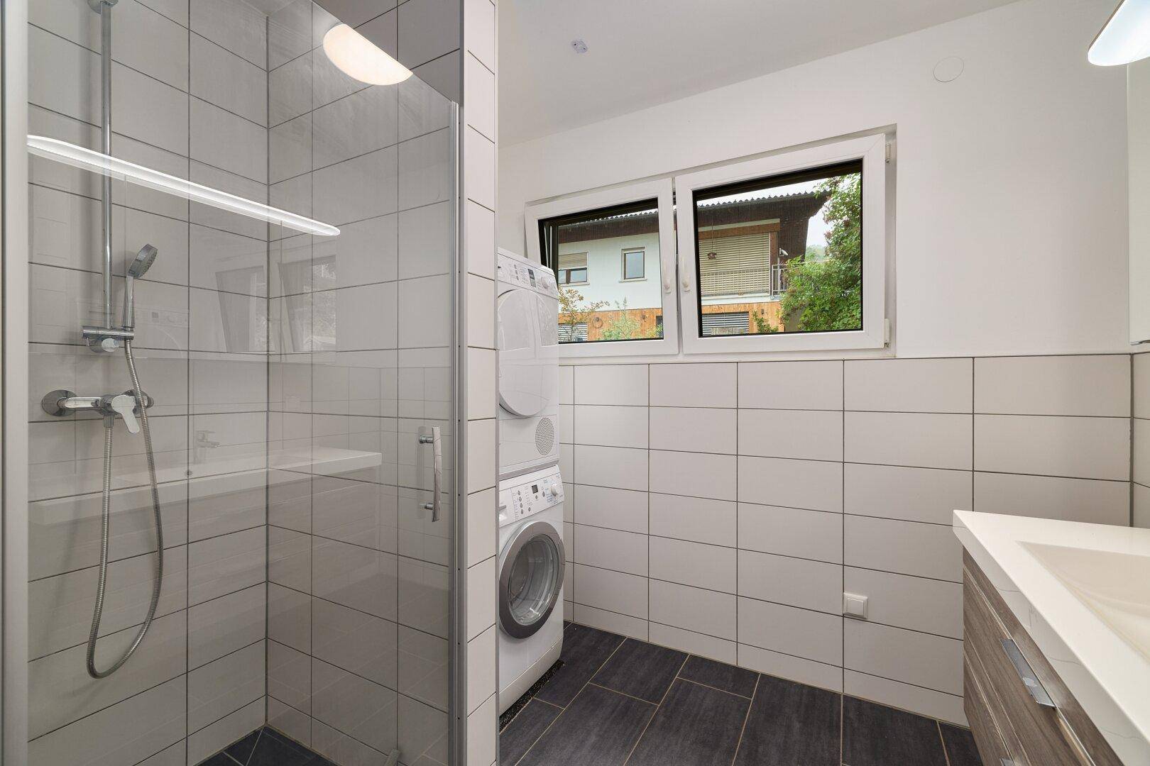 Modernes Badezimmer mit ebenerdiger Dusche, Waschmaschine und Trockner sowie einem Fenster.