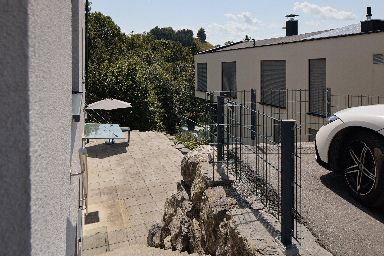 Gepflasterte Terrasse mit Sonnenschirm und Tisch, umgeben von einer Steinmauer und Grünflächen.