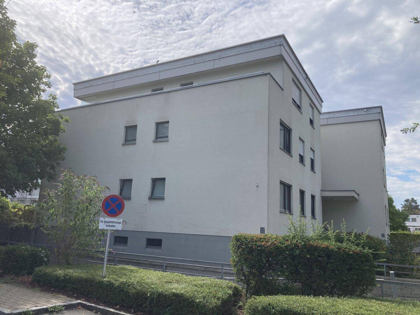 Seitenansicht eines Mehrfamilienhauses mit Fenstern, einer Rampe und gepflegten Büschen.