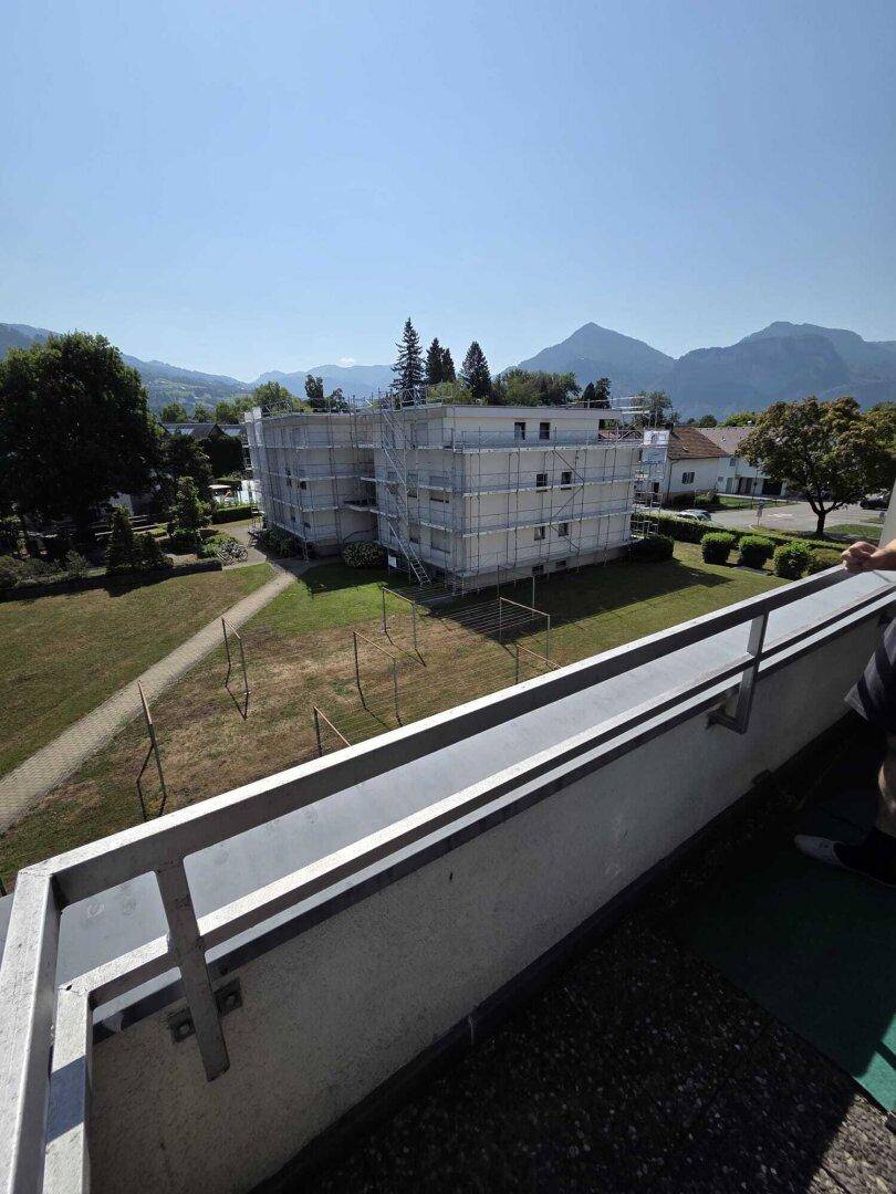 Blick vom Balkon auf ein Gebäude mit Gerüst, grüne Umgebung und Berge im Hintergrund.