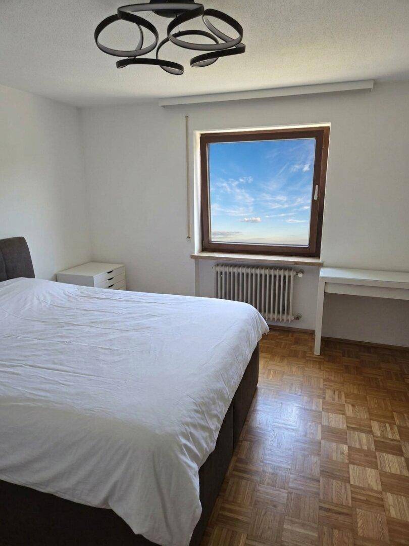 Schlafzimmer mit Bett, Parkettboden und einem Fenster, das viel Tageslicht hereinlässt.
