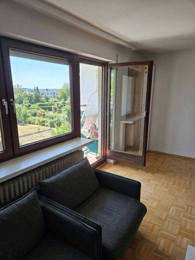 Gemütliches Wohnzimmer mit dunklem Sofa, Parkettboden und Zugang zu einem Balkon mit Grünblick.