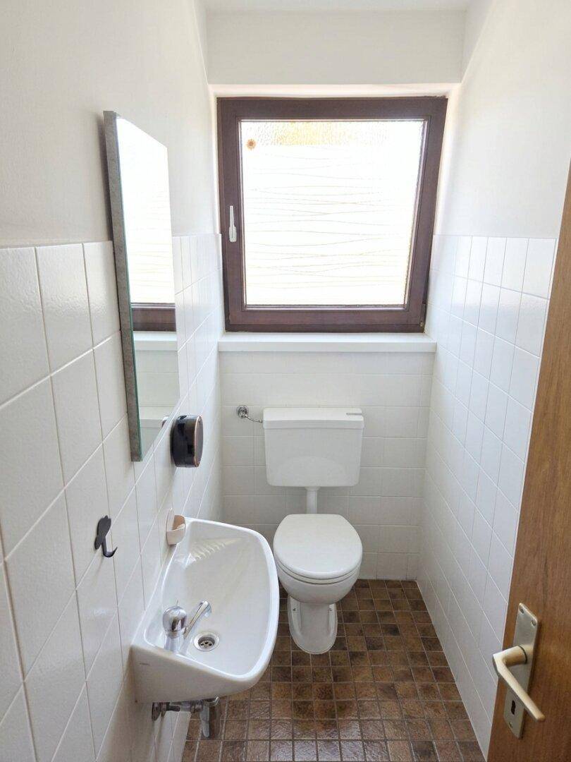 Kleines Gäste-WC mit weißen Fliesen, Waschbecken, Toilette und einem Fenster für Belüftung.