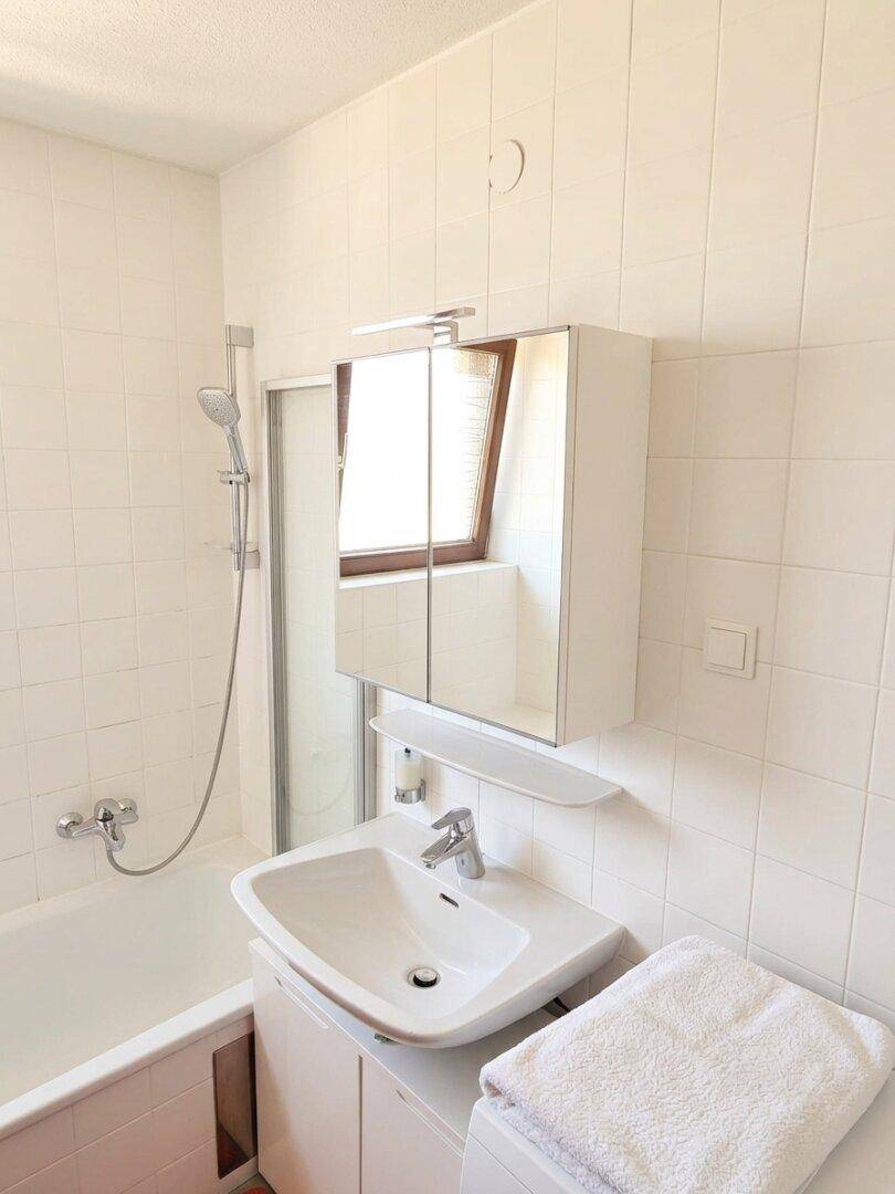 Helles Badezimmer mit weißen Fliesen, Waschbecken, Badewanne mit Duschfunktion und Spiegelschrank.