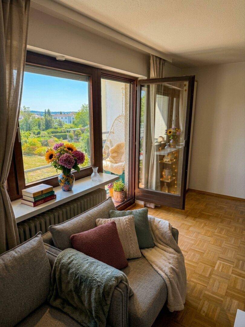 Freundliches Wohnzimmer mit Sofa, Blumen und Zugang zum Balkon mit schöner Aussicht ins Grüne.