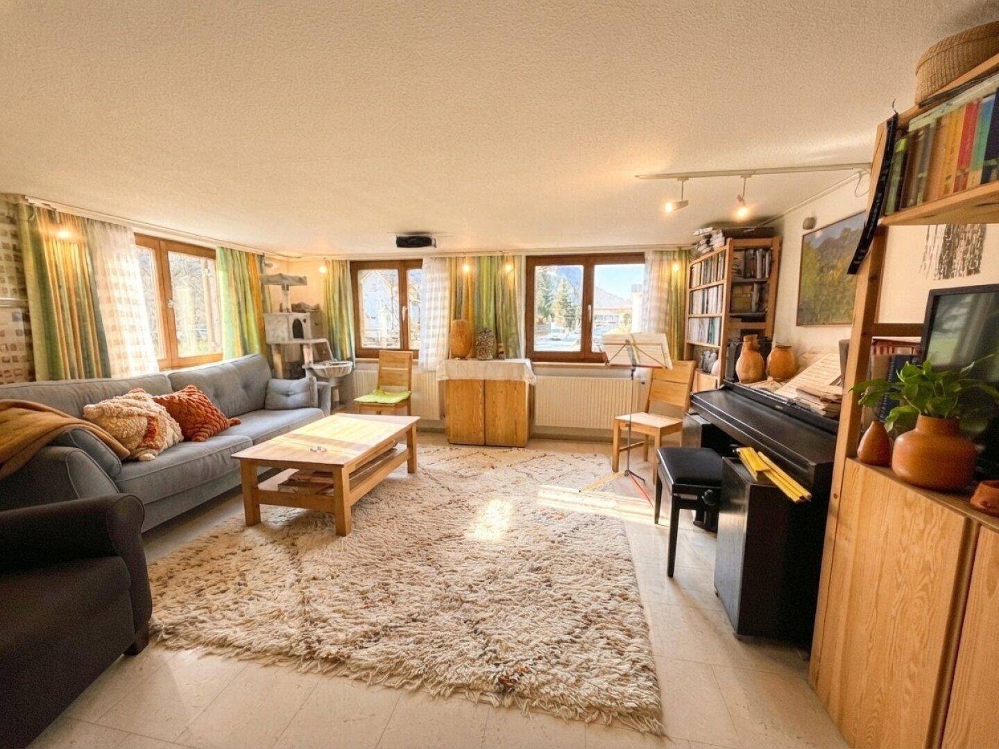 Geräumiges Wohnzimmer mit Sofa, Klavier und Bücherregalen, hell durch mehrere Fenster.
