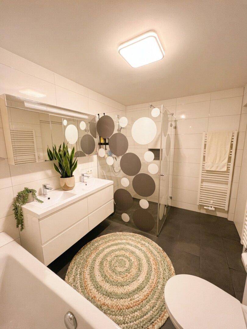 Helles Badezimmer mit Doppelwaschbecken, Dusche und Badewanne auf dunklen Fliesen.